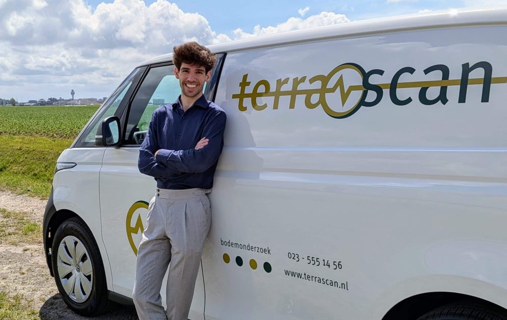 Een projectleider leunt tegen een bedrijfsbusje van een bodemonderzoeksbureau. Het busje staat geparkeerd naast een weiland.